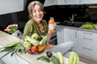 Leinwandbild Motiv Portrait of a young and cheerful woman drinking juice, sitting with healthy raw food on the kitchen at home. Vegetarianism, wellbeing and healthy lifestyle concept