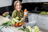 Fototapeta  - Portrait of a young and cheerful woman drinking juice, sitting with healthy raw food on the kitchen at home. Vegetarianism, wellbeing and healthy lifestyle concept