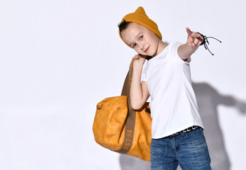 Teenage kid with orange travel bag, in hat, t-shirt, blue jeans, bracelet. Smiling, showing victory hand, posing isolated on white