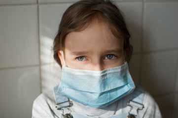 Portrait of little sad girl in a protective disposable mask stays at home because of the pandemia of Coronavirus Covid-19 in self-isolation on white tiles background on the kitchen during sunny day