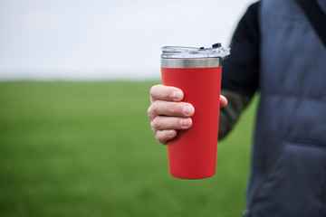 Close-up picture of male hand, holding red travel mug with hot tea coffee drink outside in the field in spring summer. Healthy lifestyle concept.