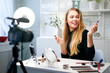 © artiemedvedev - Beauty blogger woman filming daily make-up routine tutorial near camera on tripod. Influencer blonde girl live streaming cosmetics product review in home studio. Vlogger female comparing lipsticks.