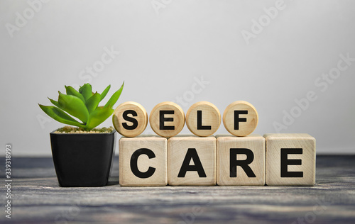 Fotomural SELF CARE - text on wooden cubes, green plant in black pot on a wooden backgroun