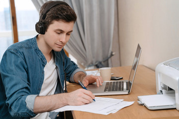 happy caucasian man focused on making notes while listening online webinar at home, wearing headset,