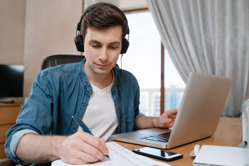 Happy young man in headphones sitting at desk in home office, writting on papers, using laptop for distance education, doing homework, writing notes, listerning audio course, watching webinar