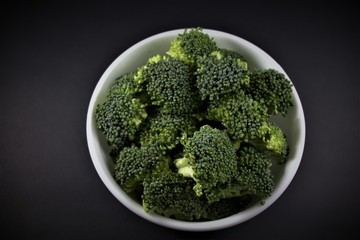 Pieces of broccoli in a white round bowl isolated on black background. Fresh and raw, green plant of cabbage Brassicaceae family, genus Brassica. Scientific name: Brassica oleracea var. italica