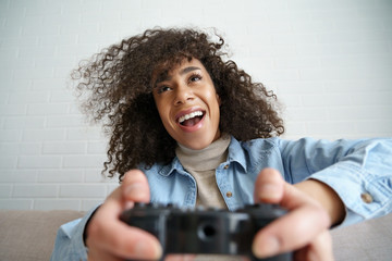 Excited young african american ethnic teen girl gamer holding gamepad joystick controller playing videogame sitting on sofa at home. Happy black woman player enjoying video gaming at home concept.