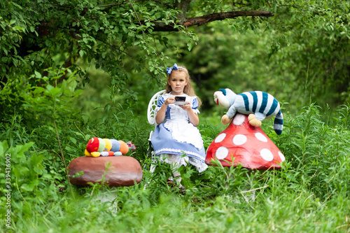 A little cute girl in the costume "Alice from Wonderland" holds a tea party at her magic table. Photographed in nature. - Buy this stock photo and explore similar images at Adobe
