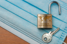 Coronavirus World Lockdown End: An Open Lock With A World Map And The Word Lockdown Engraved And A Key On A Light Blue Surgical Mask.