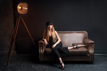 Wall Mural - Blonde model posing sitting on the couch in dark studio. Young attractive fashion model posing on dark background. Space for text