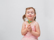 Girl On A White Background With A Lollipop On A Stick. A Girl With Ponytails In A Pink Dress With A Colorful Candy.