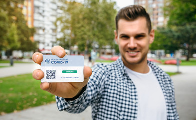 Happy young man showing health passport indicating that he is immune to coronavirus. Selective focus on card in foreground