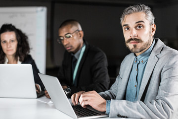 Wall Mural - Concentrated manager sitting at table and looking at camera. Focused man working with laptop in office at night. Business, working late concept