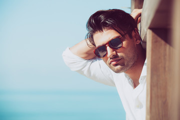 Wall Mural - Young attractive man with sunglasses looking out over the sea during the summer. He looking forward, dressed in a white shirt and leaning on a wooden construction.