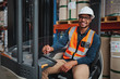 © StratfordProductions - Smiling forklift driver with spectacles sitting in vehicle in warehouse looking at camera wearing a white helmet and safety vest