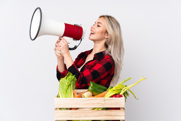 Farmer girl holding a basket full of fresh vegetables over isolated white background shouting through a megaphone