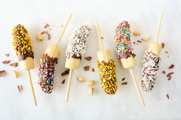 Poster - Frozen chocolate dipped banana pops with nuts, sprinkles and coconut. Top view scattered on a white marble background.