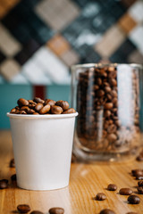 Two cups with roasted coffee beans - one transparent glass and one small paper cup on the black and brown wooden table in the cafe