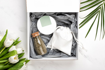 Cosmetic gift set of body or face cream container and bag with soap and a bottle of bath salts. Tulip flowers and palm leaf near box on white marble background, flat lay or top view