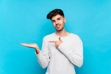 Wall Mural - Young man over isolated blue background holding copyspace imaginary on the palm to insert an ad