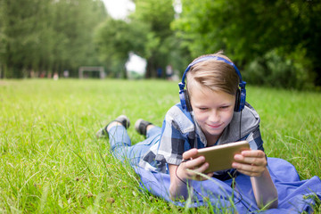 Laughing kid boy 12-16 year old with headphones outdoors. Childhood. Schoolboy. Lying on grass, summer.