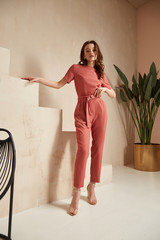 Wall Mural - Beautiful woman fashion model brunette hair tanned skin wear pink red overalls suit sandals high heels accessory bag clothes style journey safari summer collection plant flowerpot wall stairs.