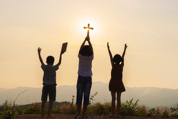 Wall Mural - Silhouette of children praying to the GOD while holding a crucifix symbol with bright sunbeam on the sky