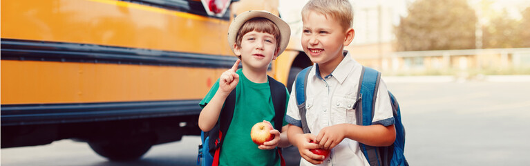 Two funny happy Caucasian boys students kids with apples standing by yellow bus on 1 September day. Education back to school. Children ready to learn and study. Web banner header for a website.