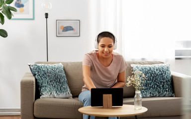 Wall Mural - people, technology and leisure concept - happy smiling young african american woman in headphones sitting on sofa and listening to music or watching movie on tablet pc computer at home