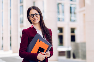 Attractive brunette  business woman in maroon suit  and glasses holding diary and and big notebook being in good mood. Business people concept.