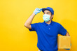 © Chanakon - Delivery man blue uniform wearing rubber gloves and mask holding parcel cardboard box on yellow background.