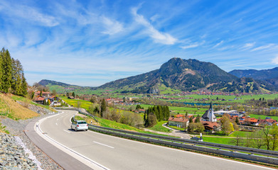 Wall Mural - Country road with car and Gruenten mountain in spring. Allgäu, Bavaria, Germany