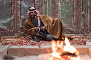 Bedouin man wearing traditional clothes praying with a tasbih while drinking tea on a carpet in front of a fire in the Saudi desert