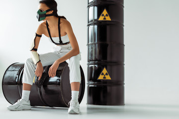 futuristic african american woman in safety mask with gun near radioactive waste barrels on white background