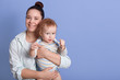 © sementsova321 - Horizontal shot of happy smiling mum holding her little son in hands, infant wearing striped bodysuit showing his fists, poses isolated on blue background. Copy space for advertisement or promotion