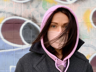 Young woman in a mask in standing in front of the painted wall, dressed in a coat and pink hood