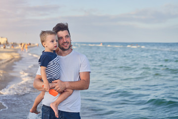 Young Caucasian dad with little son walk warm summer day along the sea coast. Summer family vacation concept. Friendship father and son.