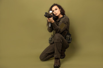 Military woman in green uniform with a vest unloading with a machine gun in her hands on a khaki background. A soldier sits with a gun