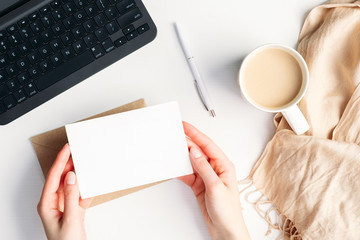 Fototapete - Female hands holding blank invitation card mockup over cozy home desk with cup of coffee, blanket, laptop computer. Hygge, autumn fall, comfort concept