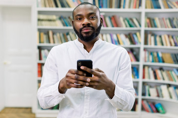 Handsome smart African American male student or businessman in white shirt writing a message or email on his mobile phone while standing in modern reading hall in library