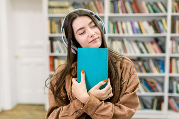 Happy young Caucasian student girl studying at the library, listening to music earphones with great pleasure and closed eyes, holding in hands favourite blue book