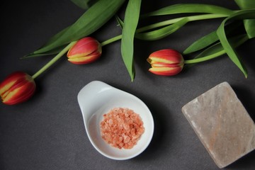 Wall Mural - Closeup of pink Himalayan salt in white bowl along with yellow red tulips and block of Himalayan salt on dark gray background. Flat lay, top view.