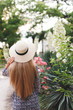 Young beautiful woman with long hair, dressed in a light blue dress with a floral print and straw boater hat, walks  in the summer on the city street near the flowering rose bushes. Back view.