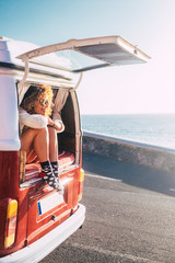 Wall Mural - Happy adult beautiful woman enjoy travel lifestyle or summer holiday vacation with old vintage van transport sit down on the back and looking the road and ocean - people free to move and live