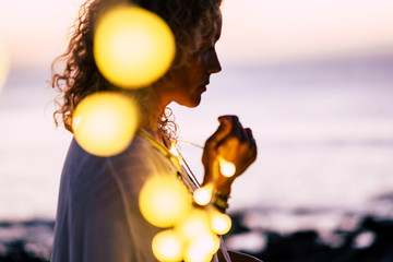 Emotional concept of hope and faith with adult woman and yellow bulb lights defocused - ocean and sunset in background - beautiful portrait for hoping conceptual image