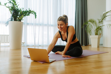 Portrait of fit young woman in sportswear watching online yoga exercises on her laptop while sitting on mat. Young woman is doing workout at home due to COVID 19 pandemic.