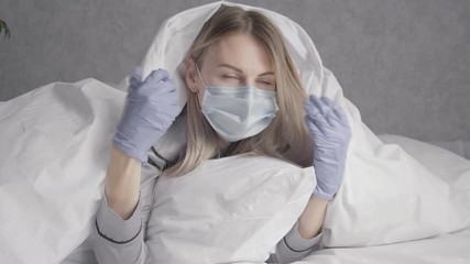 Wall Mural - a woman in a medical mask and medical gloves after reading the news about the coronavirus on the Internet hiding under a blanket in bed