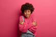 © wayhome.studio - Studio shot of good looking ethnic curly haired woman trembles from feeling cold, crosses arms over body, clenches teeth, tries to warm herself, dressed in loose knitted sweater, stands indoor