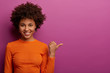 © WHstudio Leushin N - Portrait of happy dark skinned woman in bright jumper points thumb on copy space, shows advertisement of product or company banner, guarantees best quality of item, gives you recommendation.