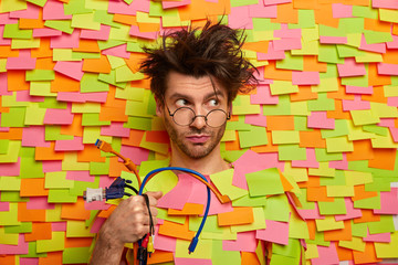 Serious thoughtful man holds bunch of colorful cables, going to repair computer, has messy hairstyle, looks through optical glasses, poses against wall with stickers. Technology, engineering